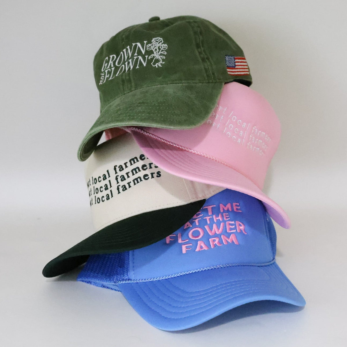 Four stacked baseball caps in green, pink, white, and blue, each with embroidered text and graphics, photographed on a plain light background.